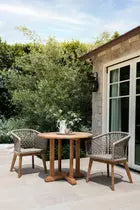 A luxurious outdoor bistro furniture setting featuring a Shelli Teak Bistro Table surrounded by stylish woven chairs, set against a serene backdrop of greenery, embodying the essence of a stylish at-home café space.