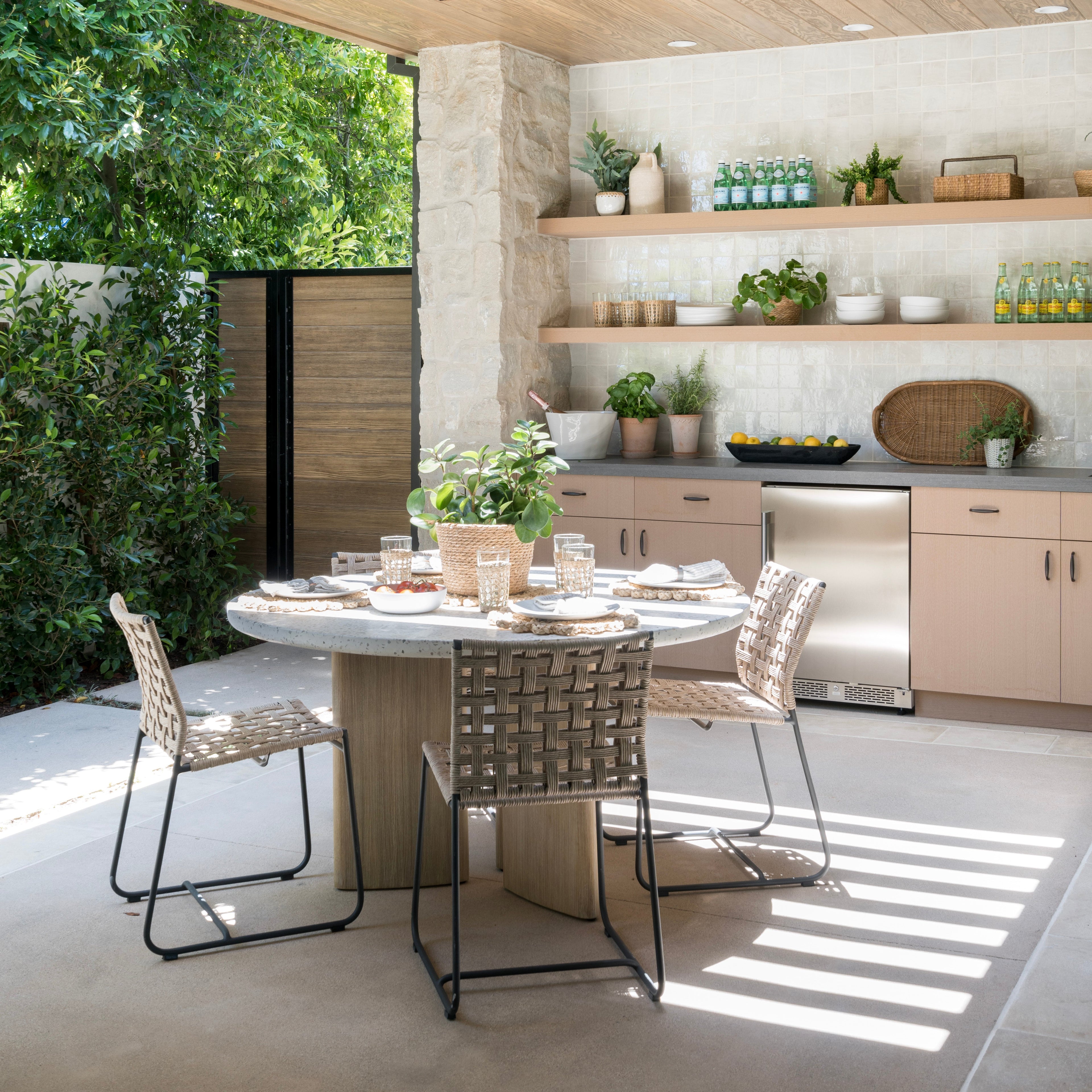 A round dining table set with glasses and plates sits on a patio surrounded by woven chairs. The background features outdoor cabinetry, shelves with plants, and bottled drinks.