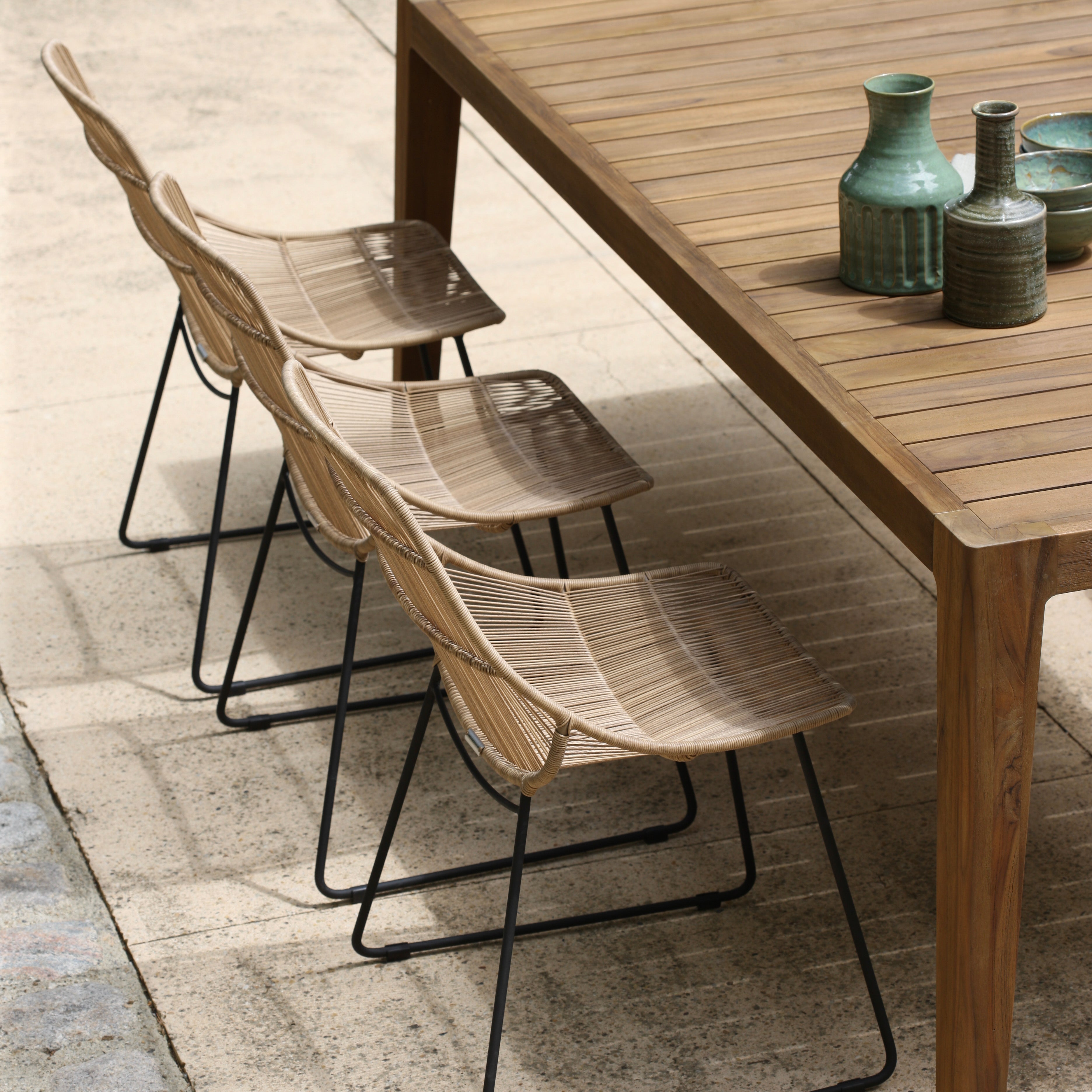 Three woven rattan chairs stand neatly beside a large wooden table with decorative ceramic vases, situated outdoors on a textured stone surface.