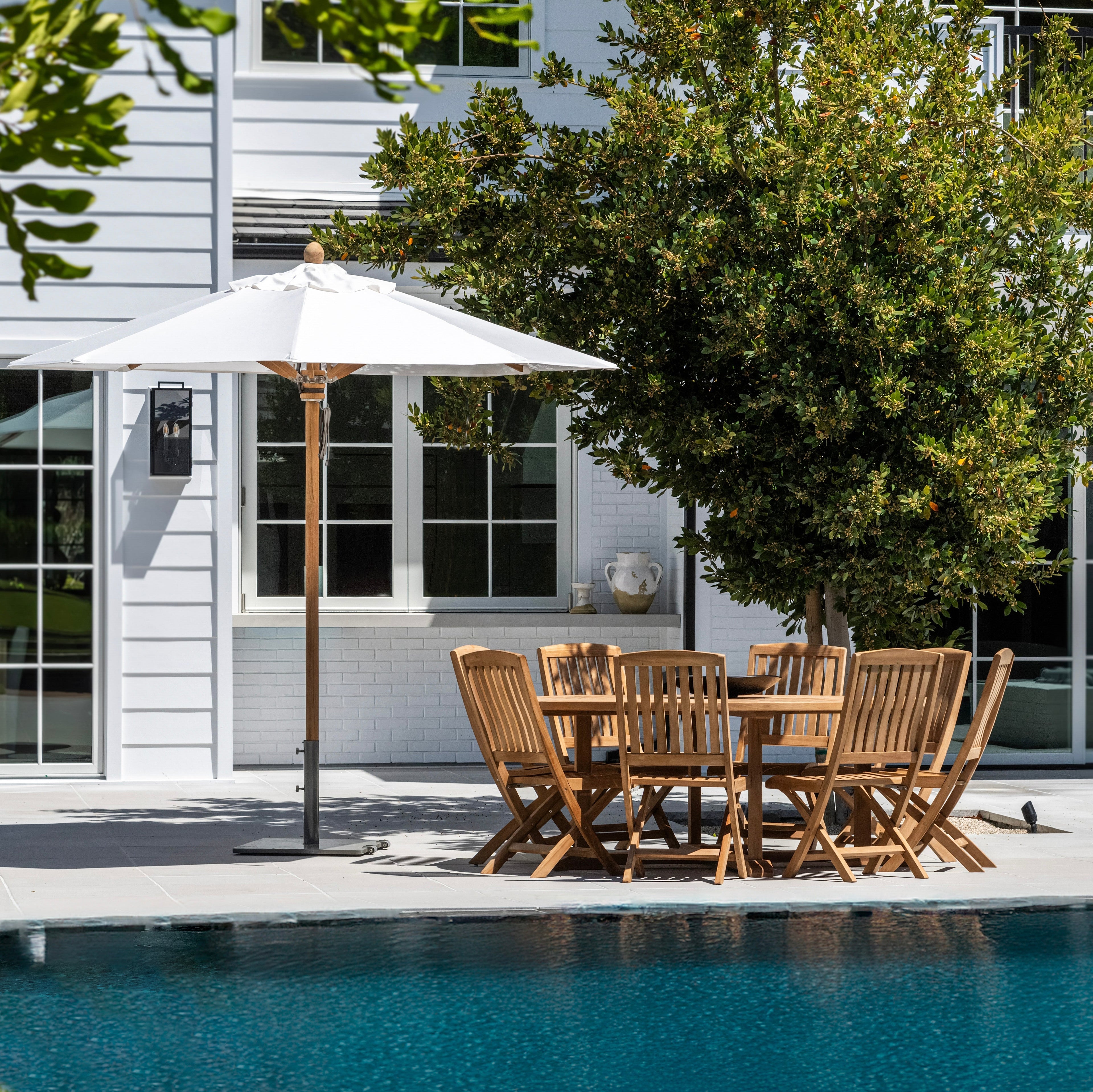 A wooden dining set with eight chairs sits under a white umbrella near a swimming pool, adjacent to a white house with large windows and greenery.