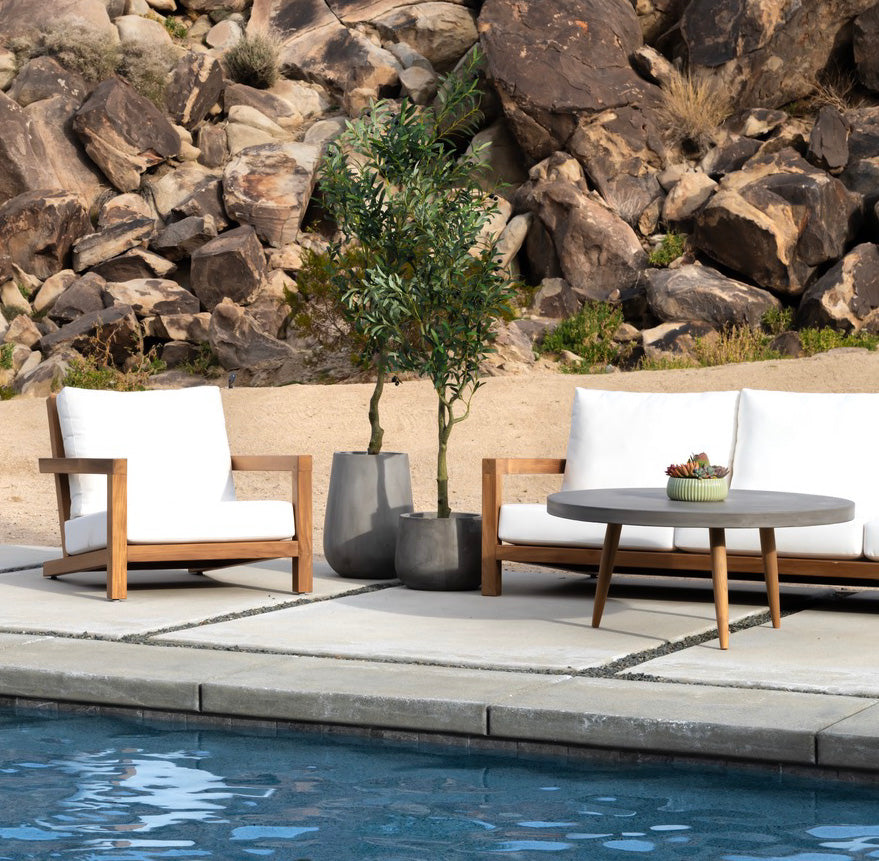 A wooden patio set with white cushions surrounds a round table, beside potted plants, on a stone patio next to a pool. Background features rocky terrain and sand.