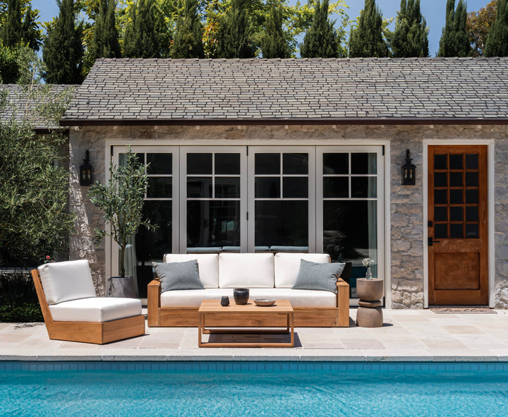 Outdoor patio furniture, including a wooden sofa with white cushions and a matching chair, rests beside a pool. A wooden table holds decor. Stone wall with greenery and windows behind.