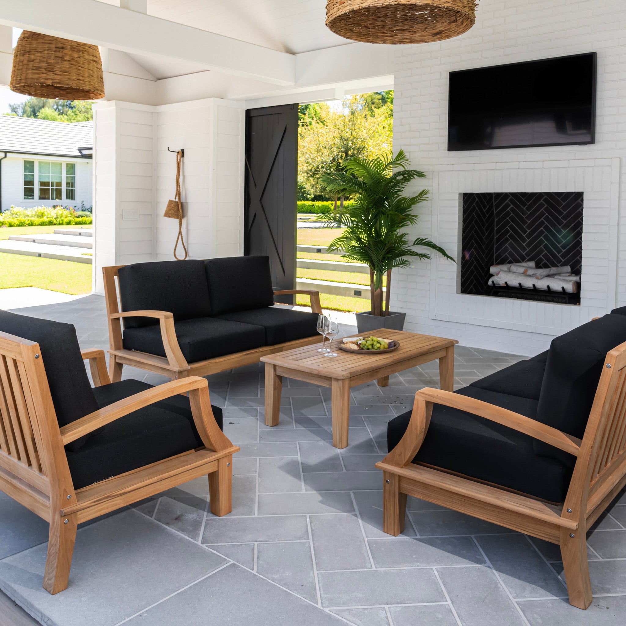 Wooden patio furniture with black cushions arranged around a low table holding grapes and wine glasses. Set on a gray-tiled floor in a covered outdoor area with fireplace, TV, and plants.