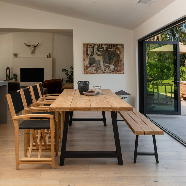 A wooden dining table is surrounded by chairs and a bench inside a modern, bright room. Art on the wall depicts a bull labeled "HAMPSHIRE." Large sliding doors reveal a lush garden.