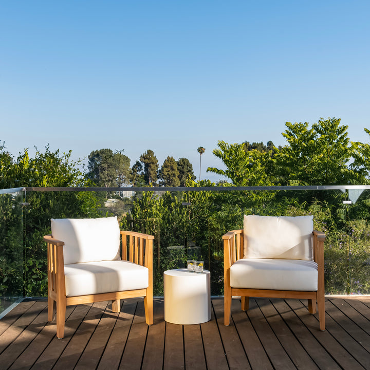 Two wooden chairs with white cushions are stationary on a wooden deck, flanking a small round table with glasses. Lush greenery and a clear blue sky create a serene backdrop.