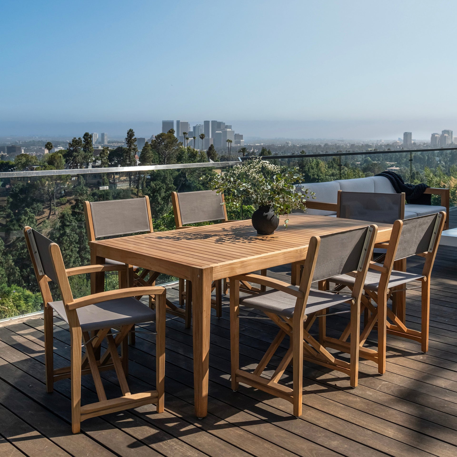 A wooden outdoor dining table is surrounded by six chairs, with a potted plant centerpiece. It sits on a deck overlooking a cityscape with trees and buildings in the distance.