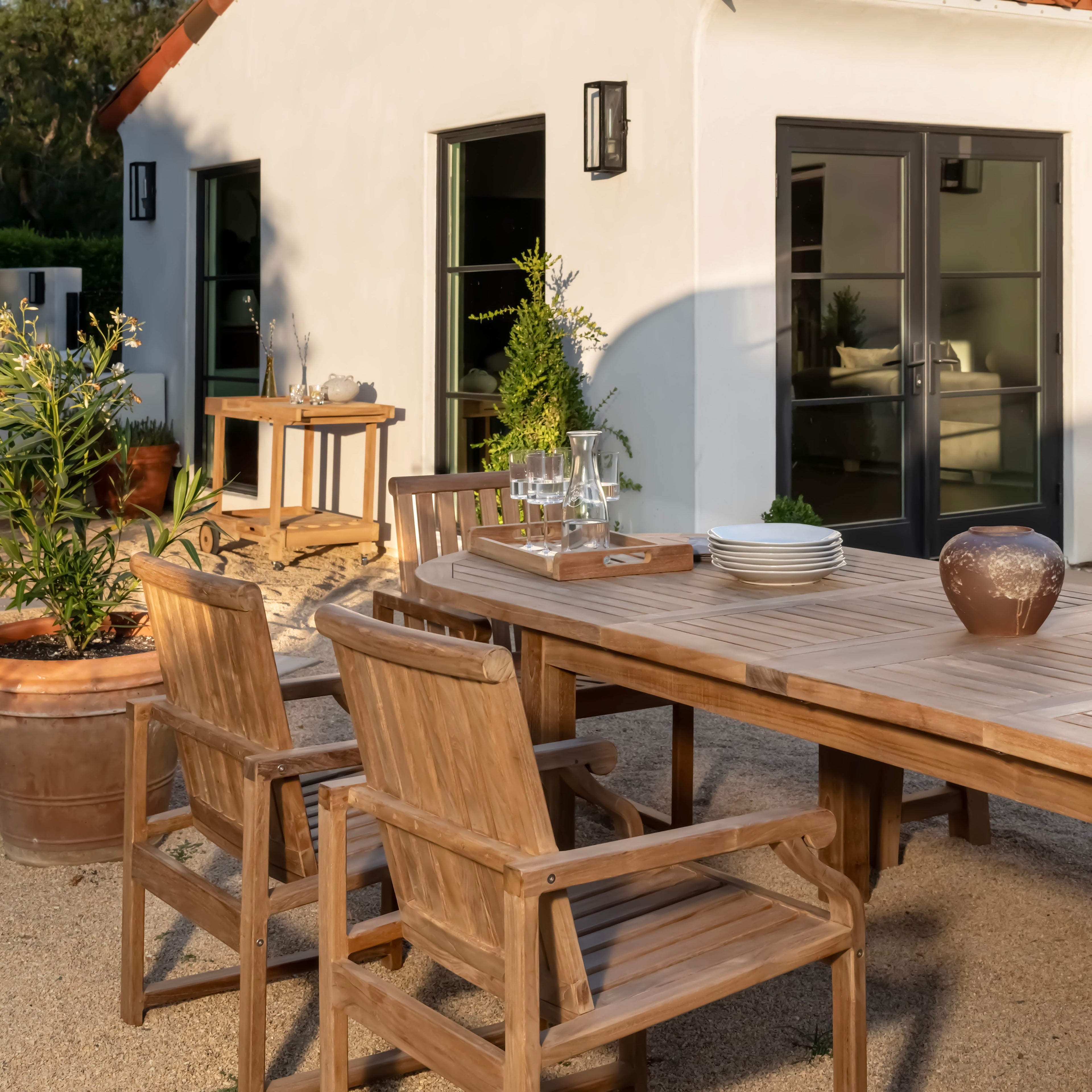 Wooden outdoor table and chairs set with glasses, plates, and a jug on it, sitting in a sunlit patio area with potted plants and a house in the background.