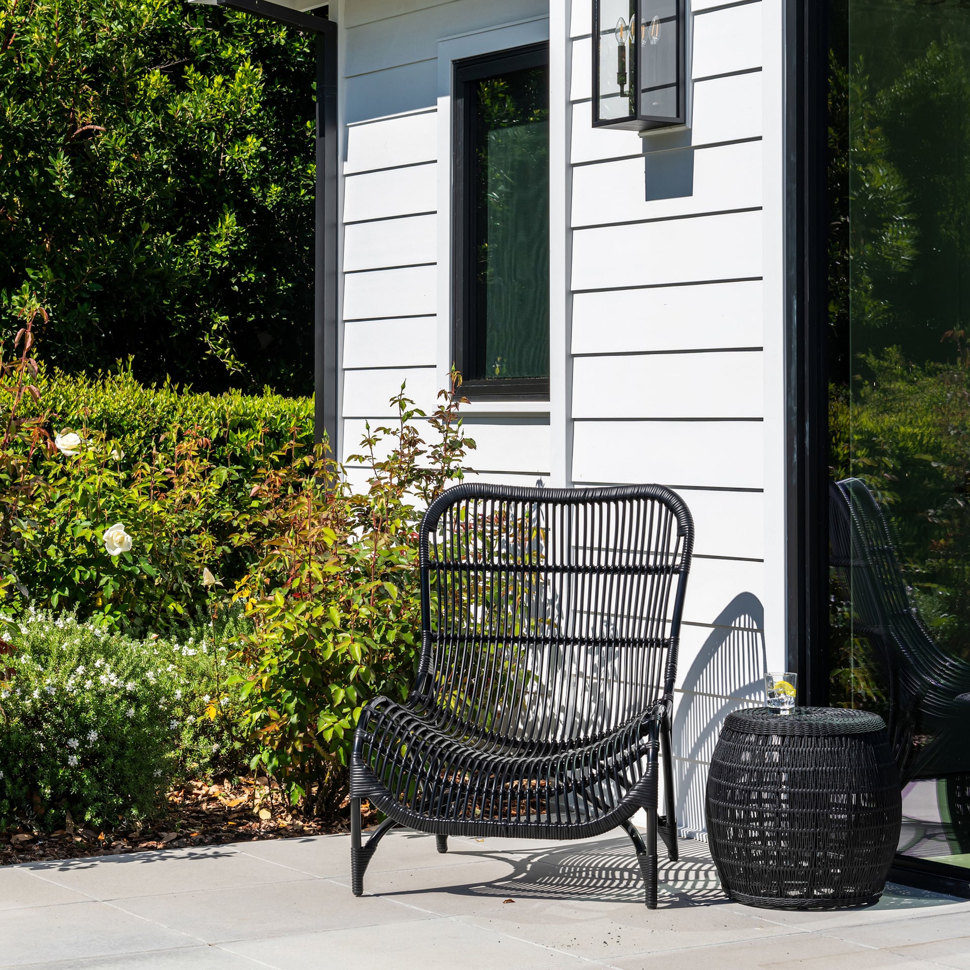 A black rattan chair and round side table sit on a patio. The sunny setting features a white-paneled house exterior, green bushes, and blooming white flowers nearby.