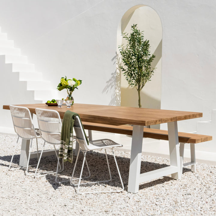 A wooden dining table with white metal chairs sits on a gravel surface, adorned with a vase of green flowers. Nearby, an olive tree is framed by an arched, white wall.