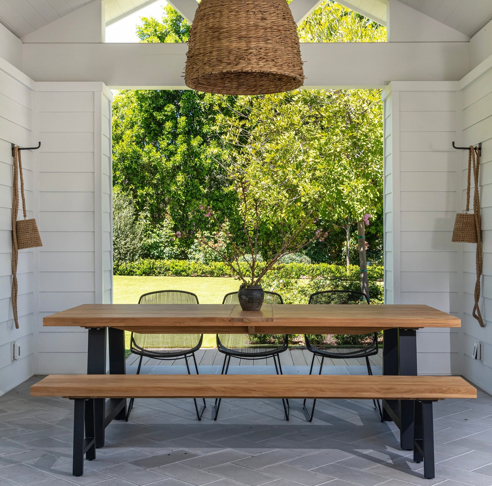 A wooden table with a bench and four chairs sits under a wicker pendant light in a patio, framed by shiplap walls and overlooking a lush garden.