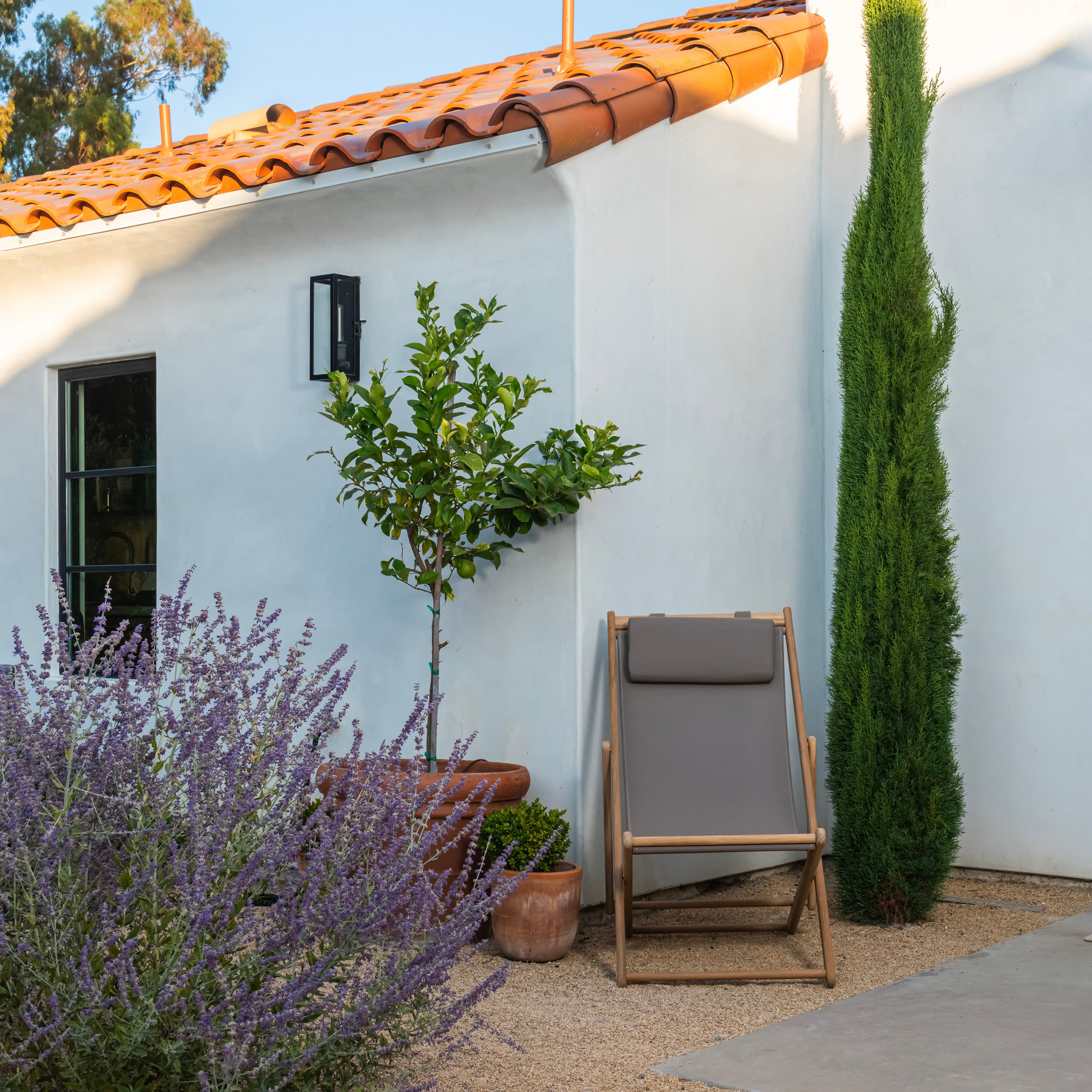 A folding chair rests on a gravel patio beside a white stucco building with a terracotta roof, surrounded by lavender, potted plants, and a tall cypress tree.