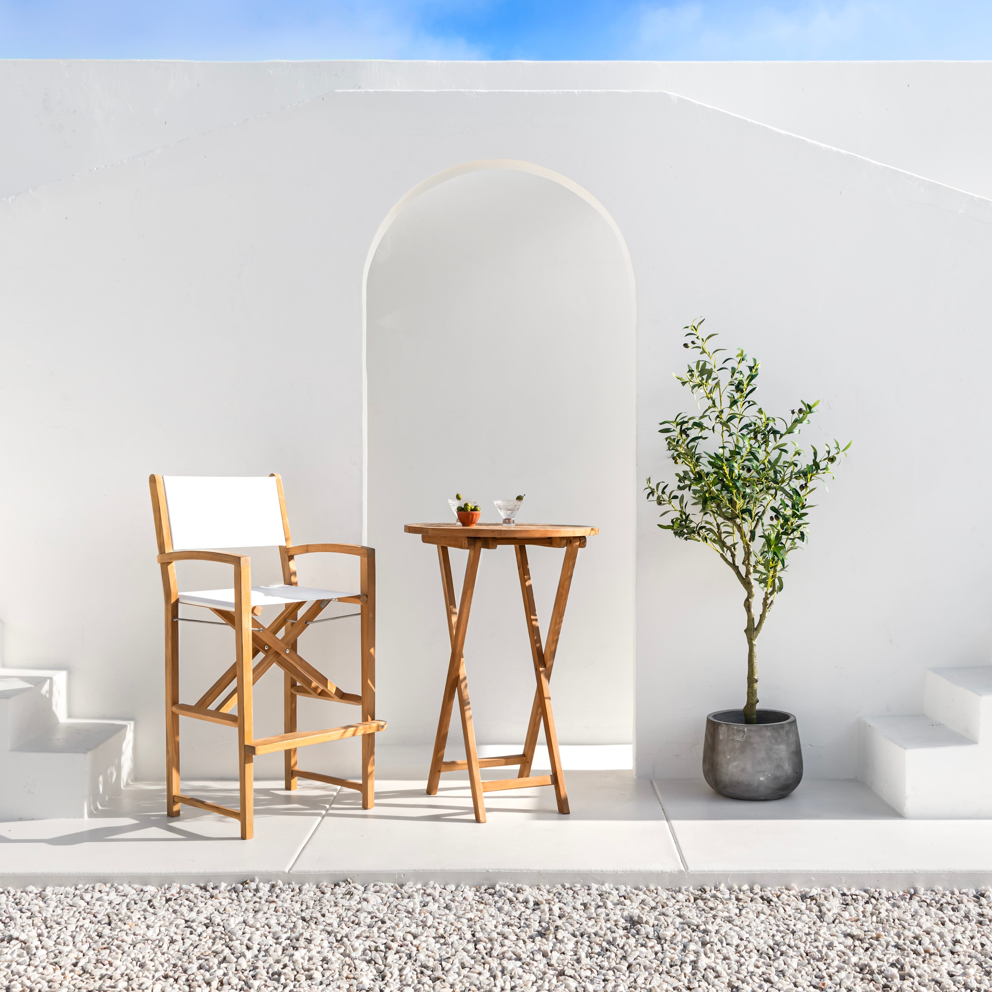 A tall wooden chair and table stand on a white patio beside an archway, accompanied by a potted plant against a smooth, white wall. Stones cover the ground nearby.