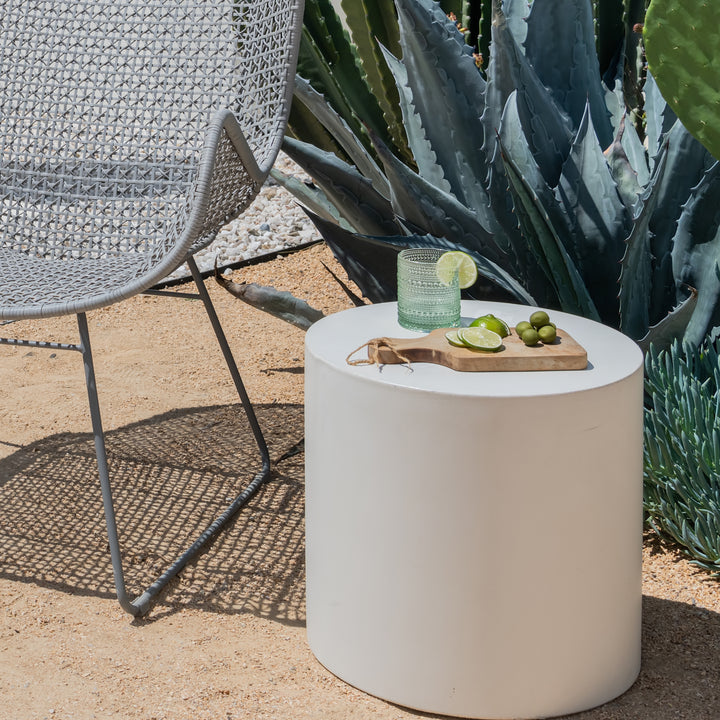 A woven chair sits beside a cylindrical white table. On the table, a glass with lime slices and a cutting board with limes are set. It's surrounded by large succulents.