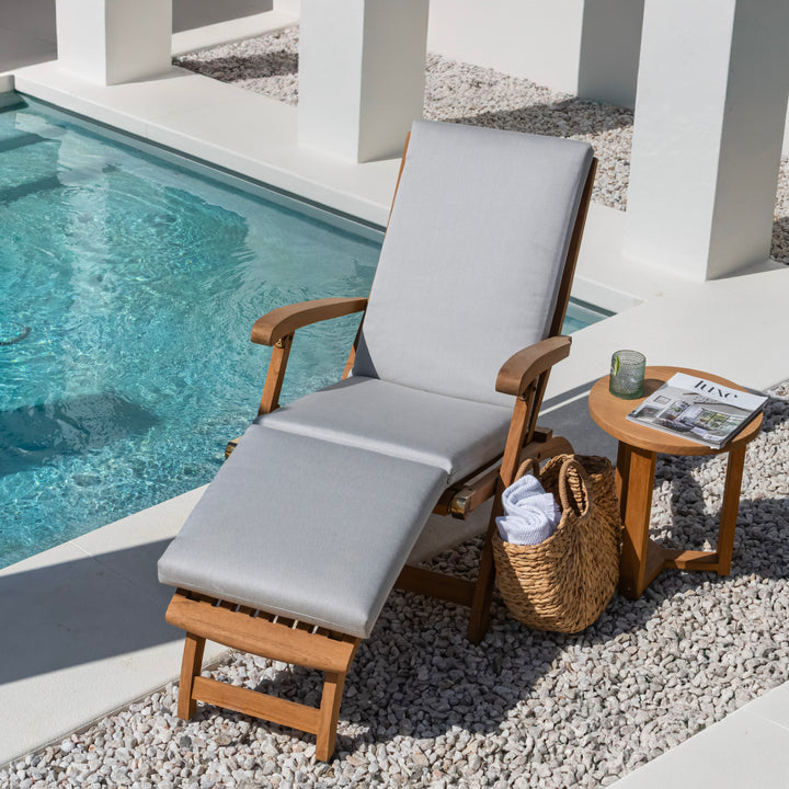 A cushioned wooden lounge chair sits by a pool, accompanied by a small table holding a drink and a magazine titled "FLANEUR," with a woven basket below on pebbled ground.