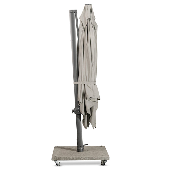 A closed, gray patio umbrella stands upright on a wheeled rectangular stone base in a gray, sparse indoor setting.