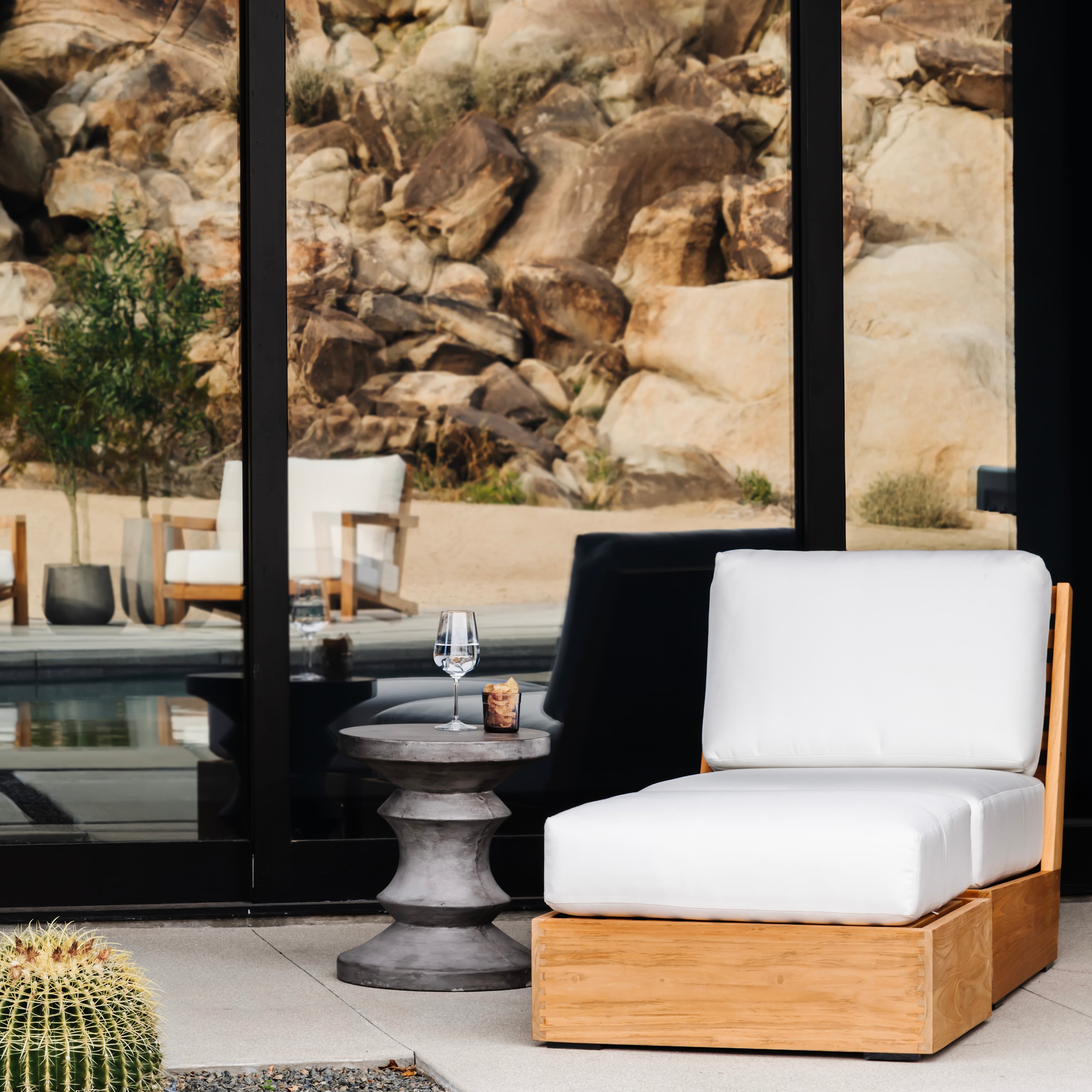 A cushioned wooden chair sits beside a modern, small table with a wine glass and candle. Reflections in glass doors show rocky terrain and distant patio furniture. Cactus in foreground.