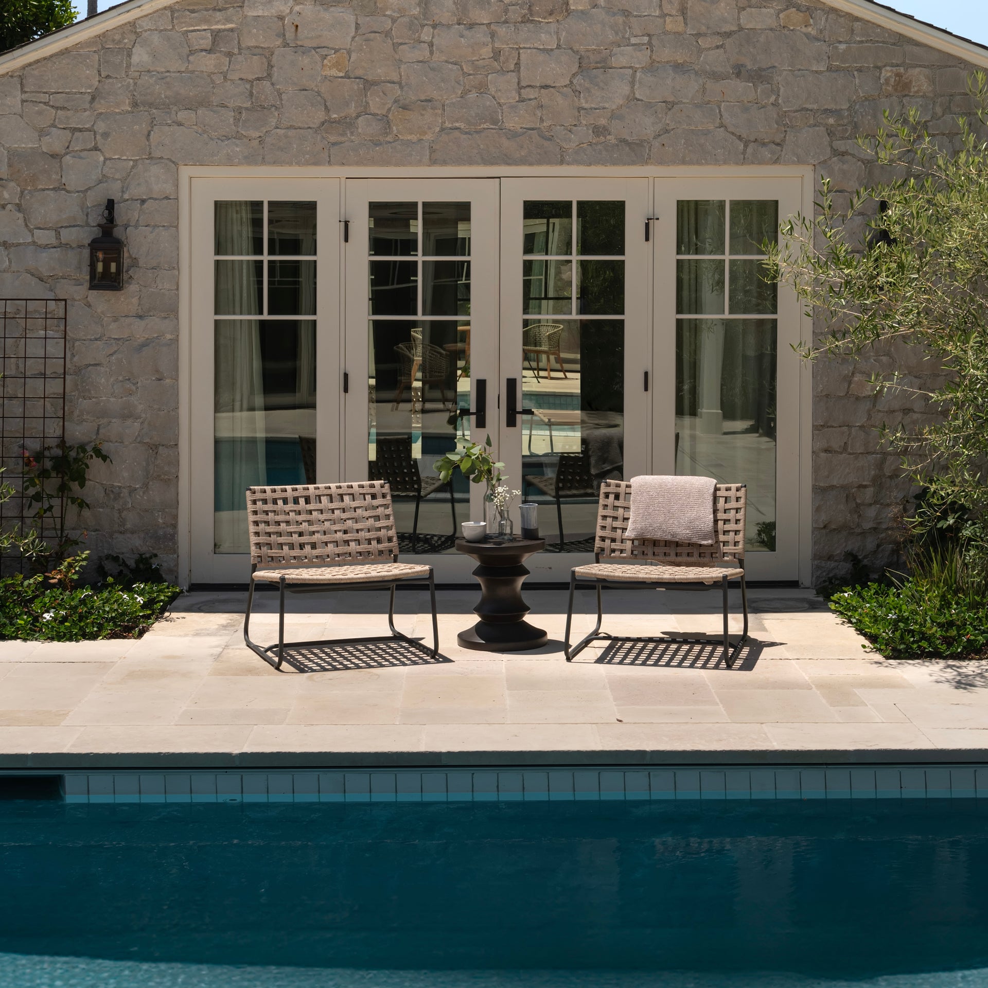 Two woven chairs face a small table holding flowers, set on a stone patio beside a clear pool. In the background, French doors open to reveal an interior space.