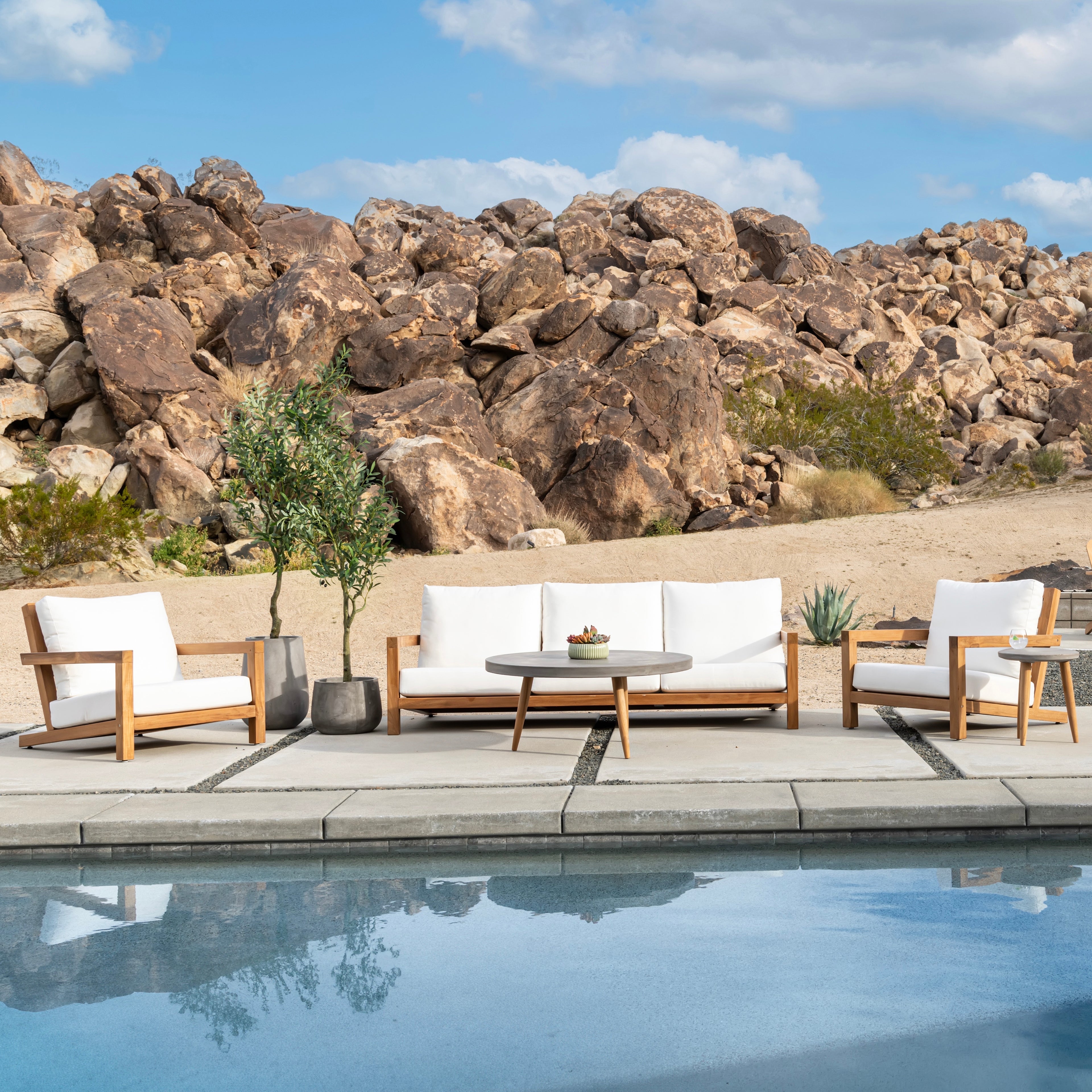 Outdoor furniture set with white cushions is arranged by a pool, featuring a couch, two chairs, and a round coffee table, against a rocky desert landscape and blue sky.