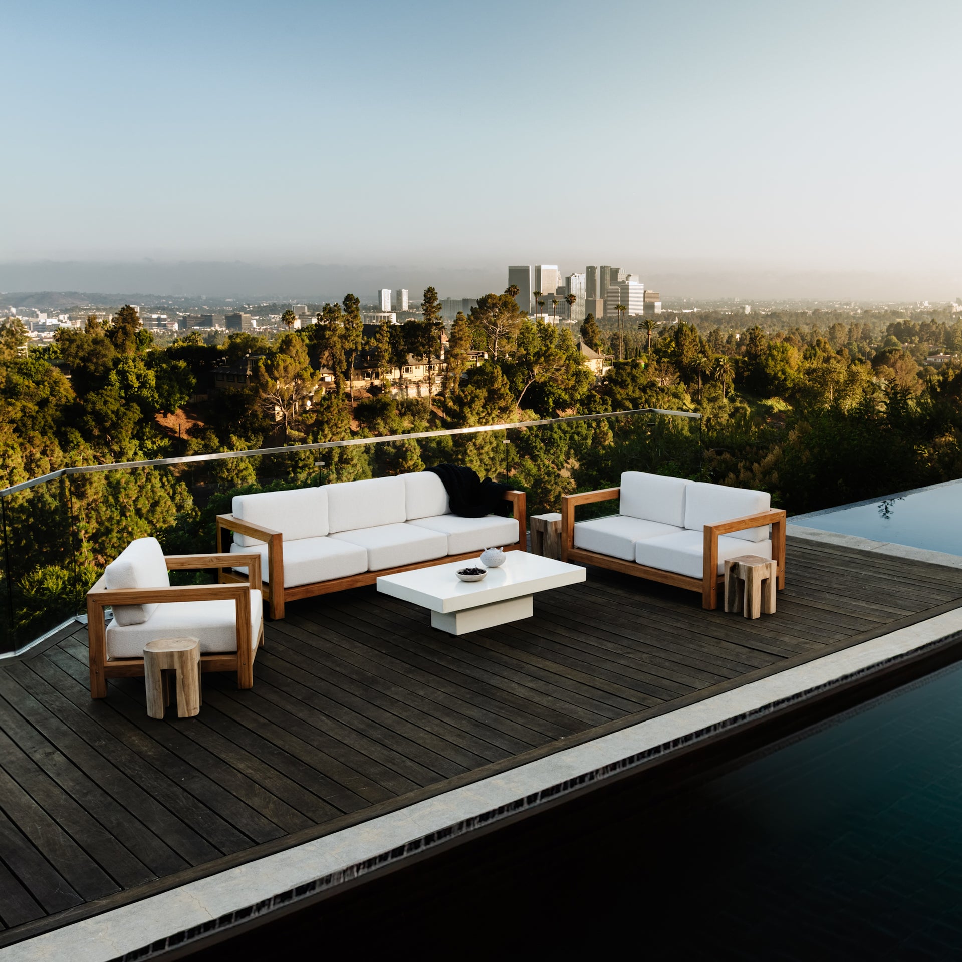 Outdoor seating area with white-cushioned wooden sofas and a central low table, set on a wooden deck. Overlooks a lush, green landscape and a distant city skyline under a clear sky.