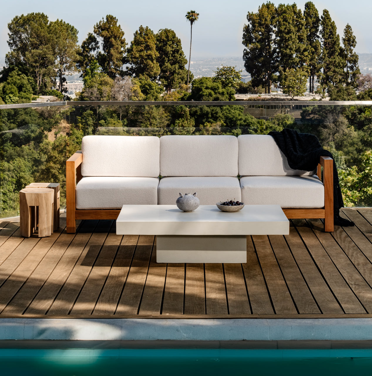 A white cushioned sofa rests on a wooden deck beside a pool, accompanied by a small square table and a flat coffee table holding two decorative items, overlooking a lush, tree-filled landscape.