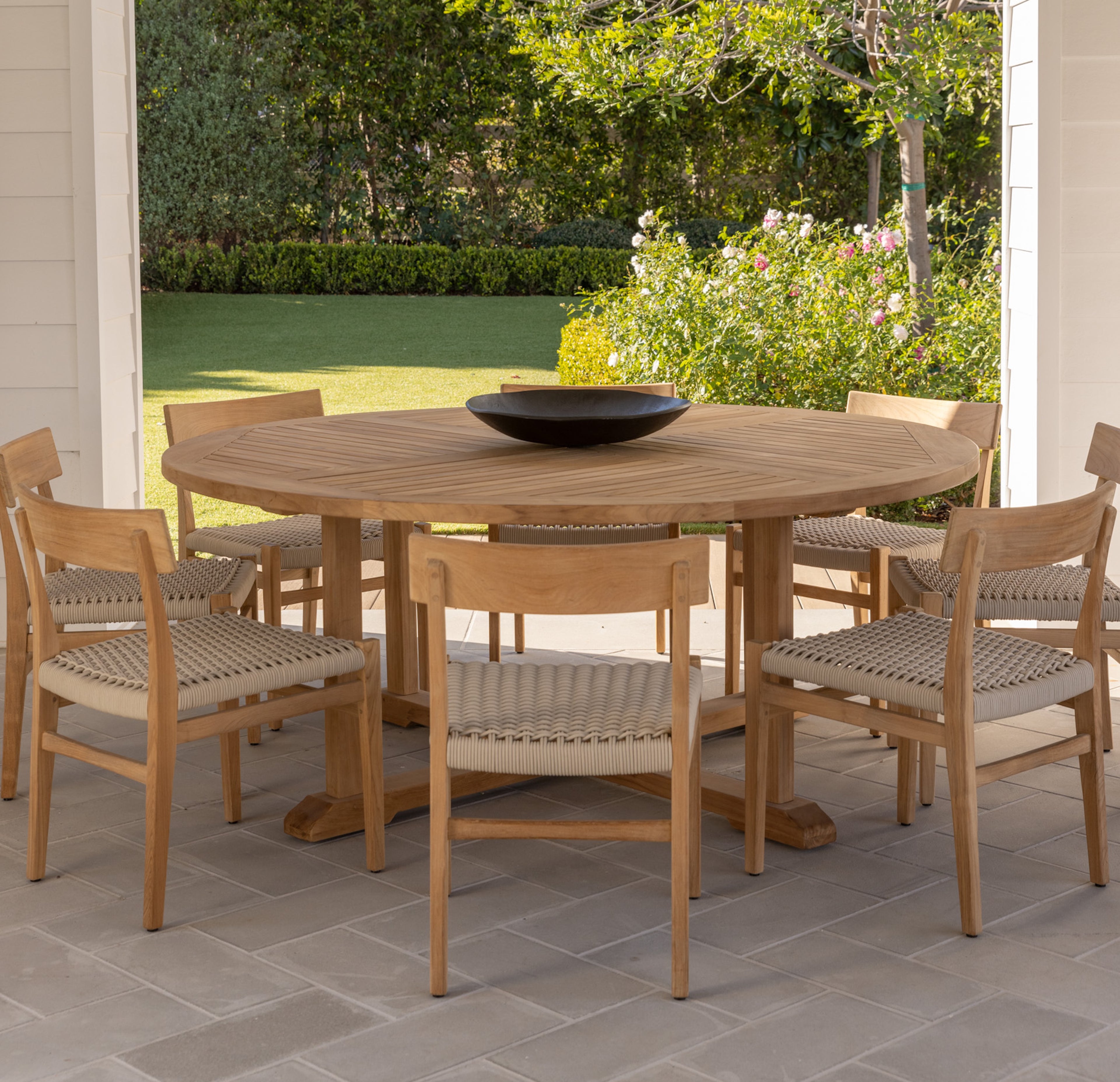 A round wooden table holds a black bowl, surrounded by six chairs with woven seats. The scene is set on a patio overlooking a manicured garden with bushes and trees.