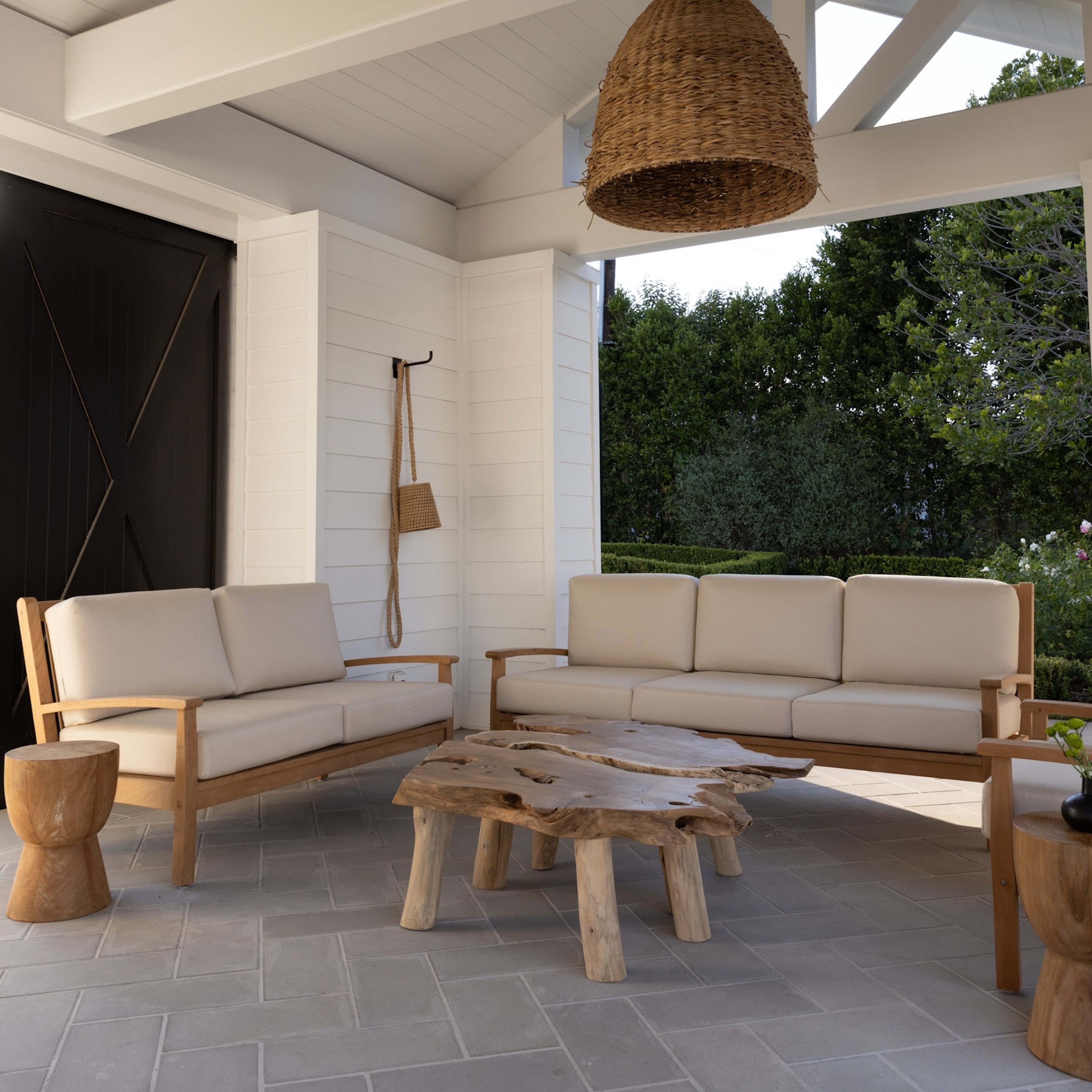 Wooden outdoor furniture set arranged on a tiled patio, including two cushioned sofas around a rustic, irregular-shaped coffee table, beneath a large wicker pendant light, surrounded by greenery.