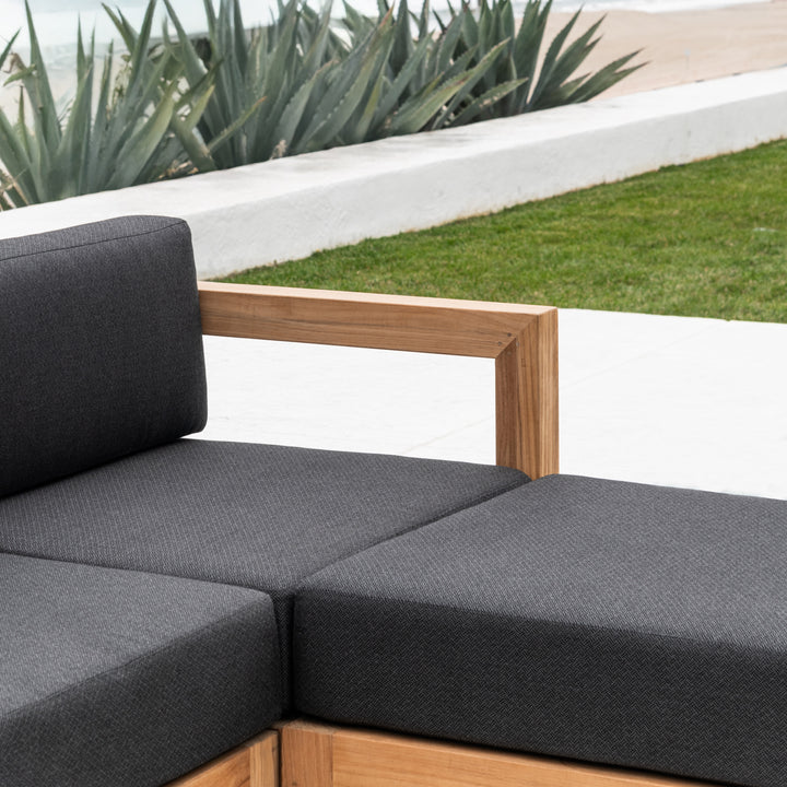 A modern outdoor sofa features dark gray cushions and wooden armrests. It is situated on a patio beside green grass and agave plants, with a white wall and walkway.