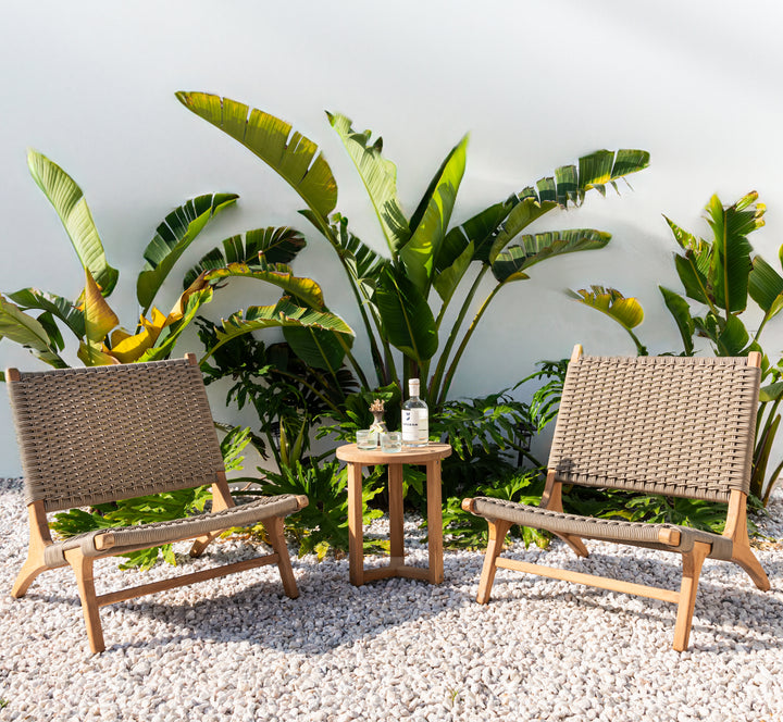 Two wicker chairs flank a small round wooden table holding a bottle, glass, and plant, set on white gravel. Large green tropical plants create a lush backdrop against a smooth wall.