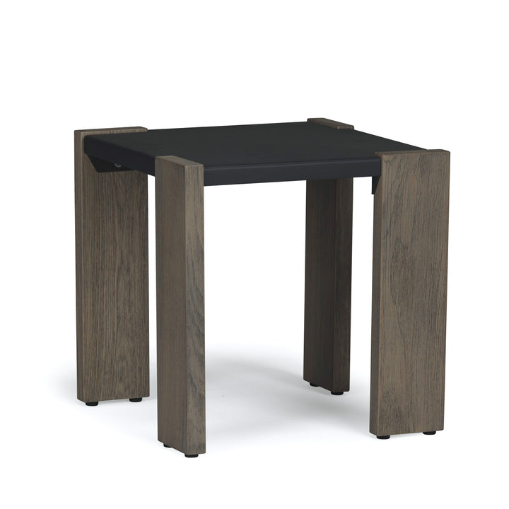 A square, black tabletop rests on four vertical, natural wood legs with minimalistic design; the table stands on a flat, neutral-colored surface.