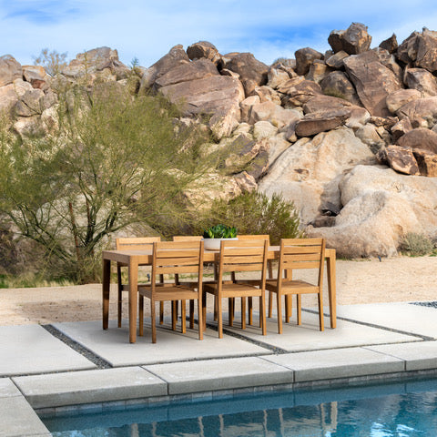 A wooden dining set stands on a patio beside a swimming pool. Rocky hills and green shrubs provide a natural desert backdrop under a blue sky.