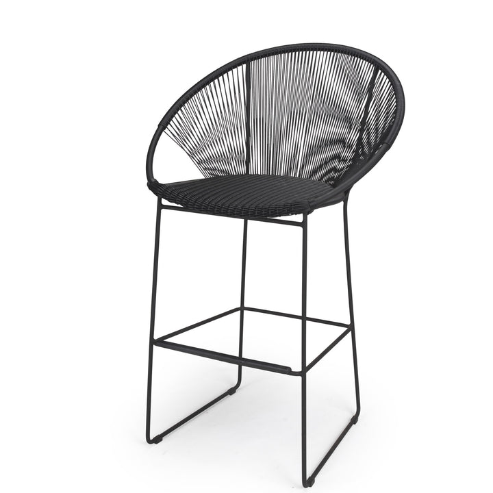 A black, round-backed bar stool with a woven design sits on thin metal legs, placed in a minimal, plain setting.