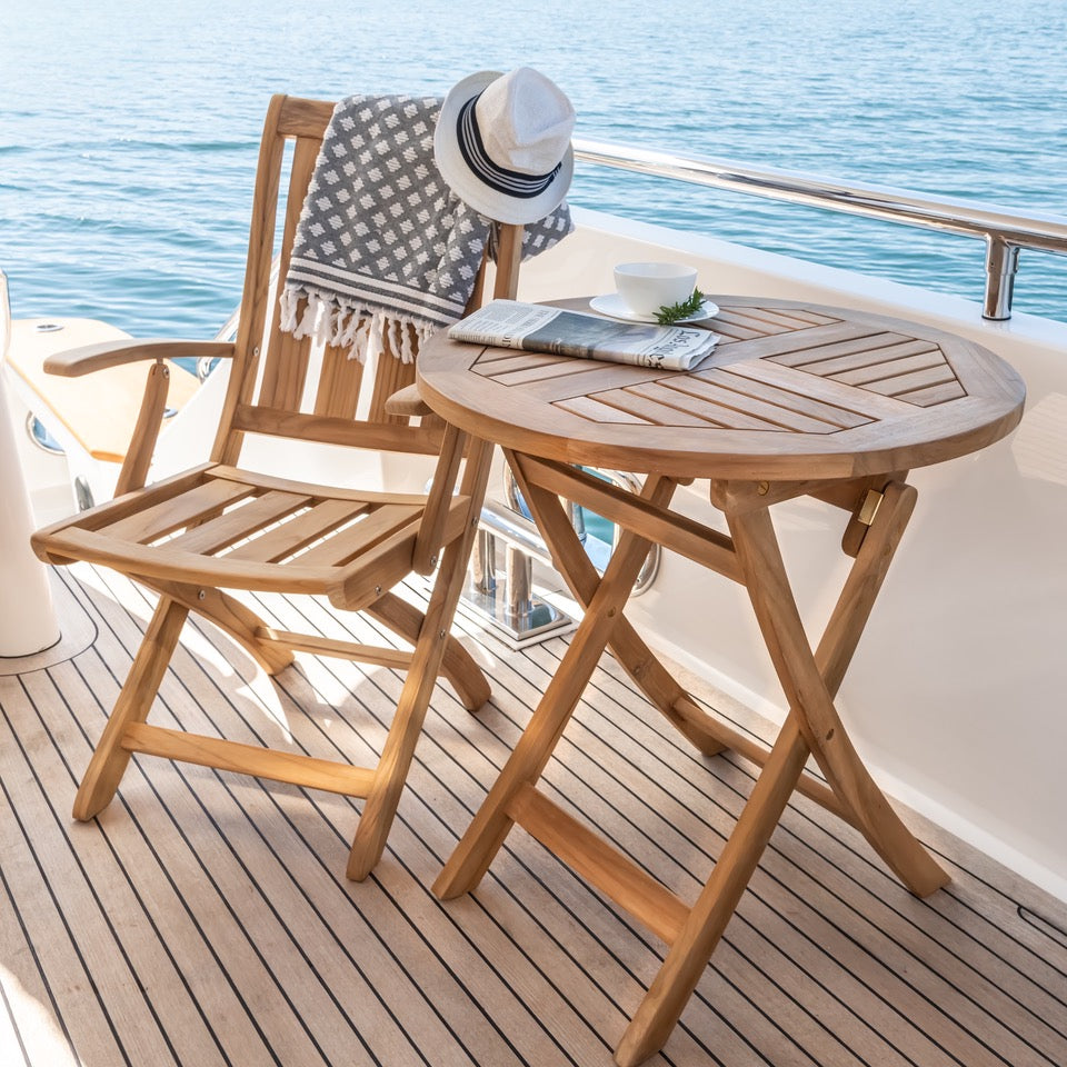 A wooden chair holds a hat and checkered blanket. A nearby table has a cup, plant, and newspaper. All are situated on a boat deck overlooking the serene blue ocean.