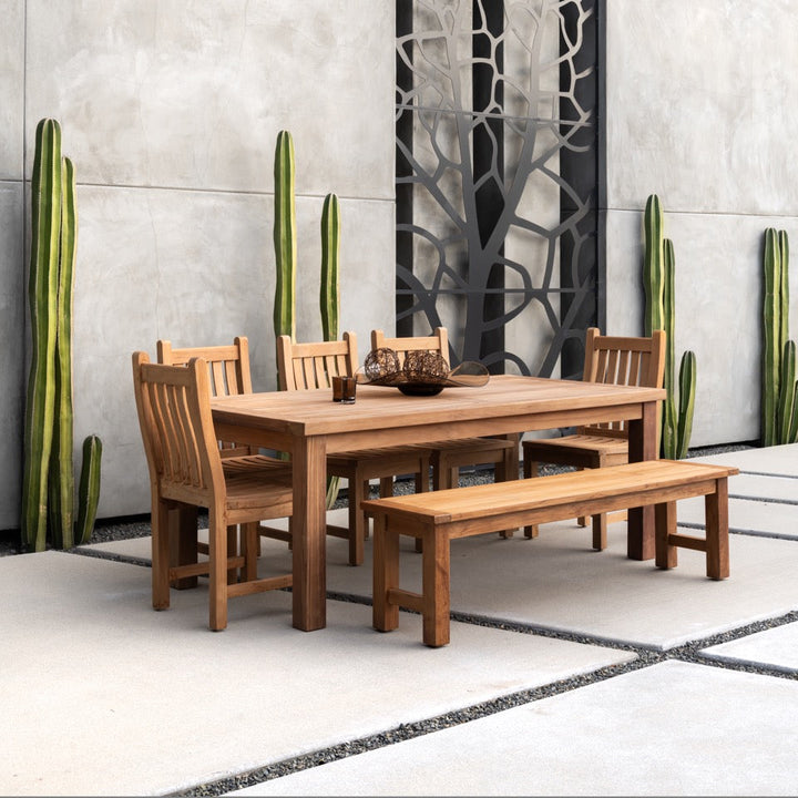 Wooden table and chairs are set on a patio with a modern concrete floor. Tall, slender cacti line the walls, while a decorative panel adds artistic interest in the background.