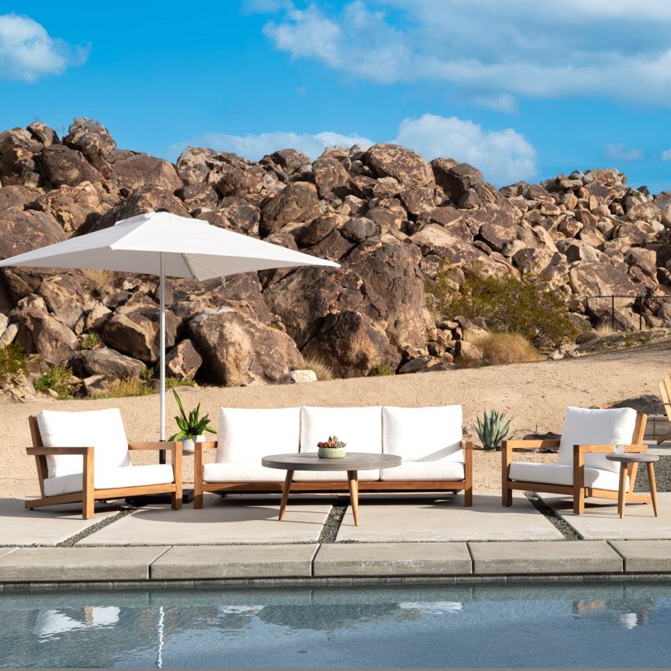 Outdoor lounge furniture sits on a poolside patio. The setup includes cushioned chairs, a white umbrella, and a round table with a plant. Rocky landscape and clear sky in the background.