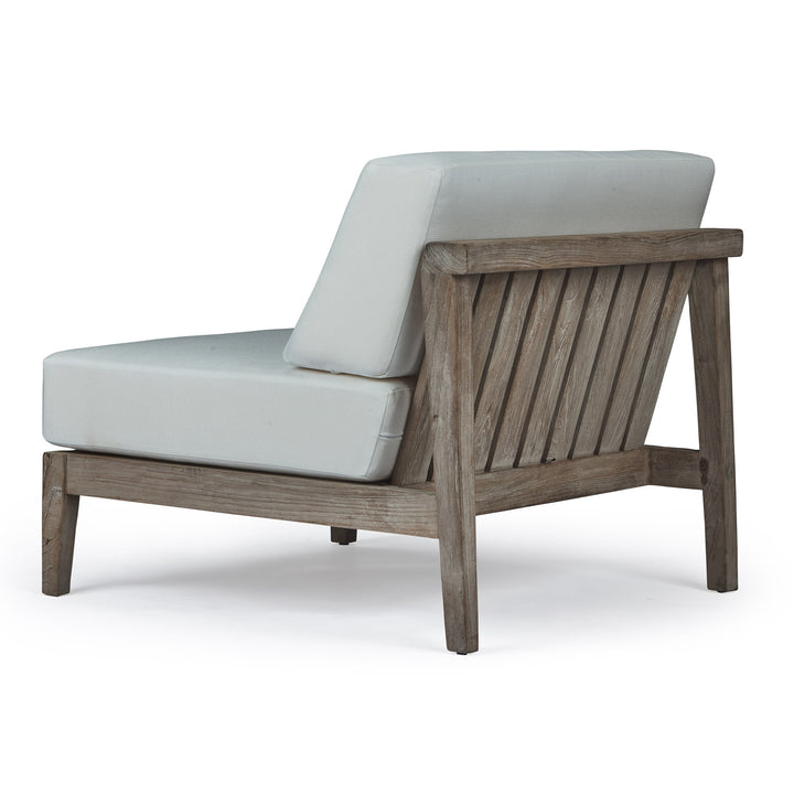 A wooden lounge chair features angled, slatted backrest and light gray cushions. It is placed in an isolated, neutral setting, highlighting its rustic, minimalist design.