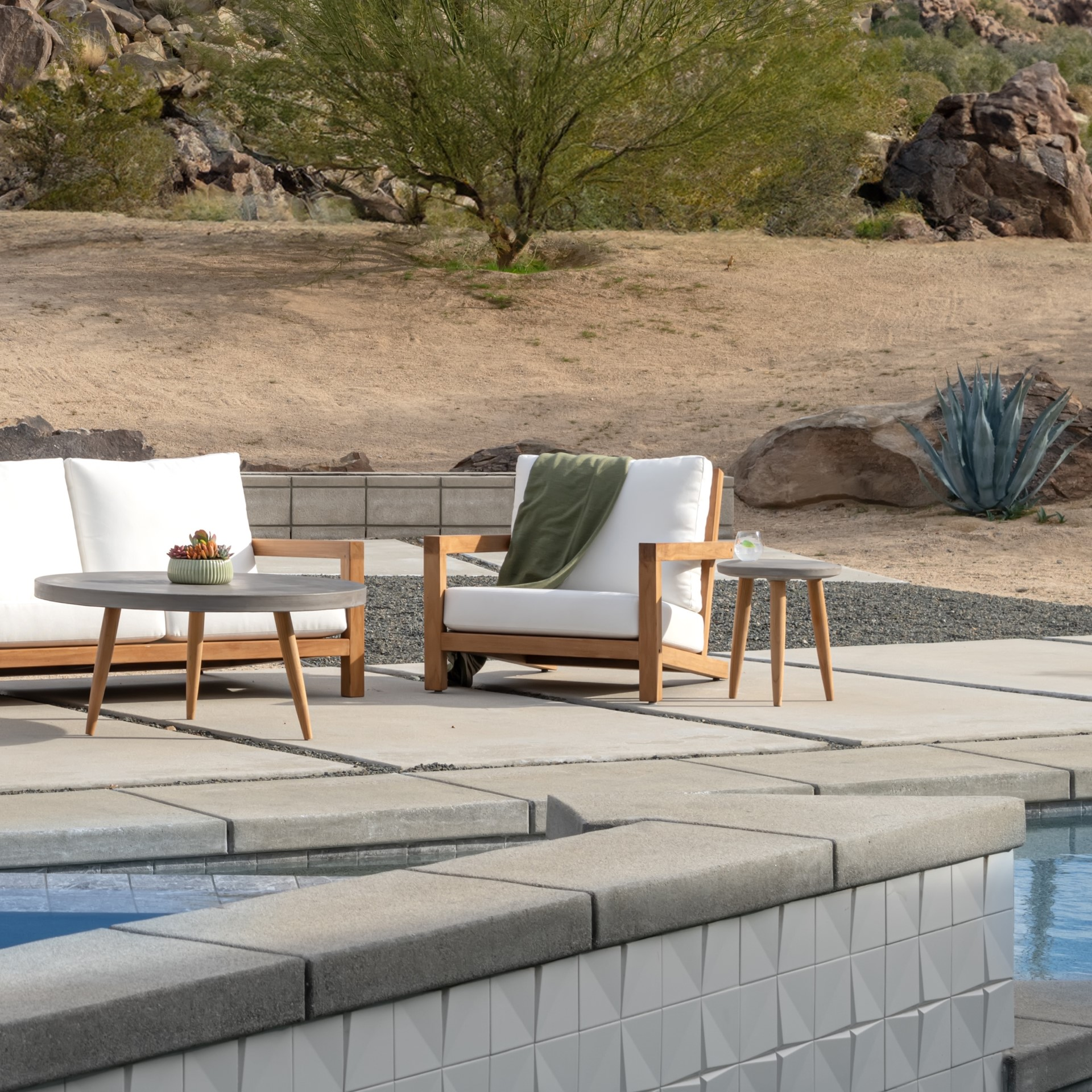 Poolside patio features white cushioned wooden chairs and tables, one holding a plant, beside a serene pool. Desert landscape with rocks, sand, and sparse greenery fills the background.