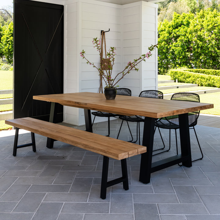 A wooden table with a floral centerpiece is flanked by a bench and chairs on a tiled patio, surrounded by greenery and a partially open black door.