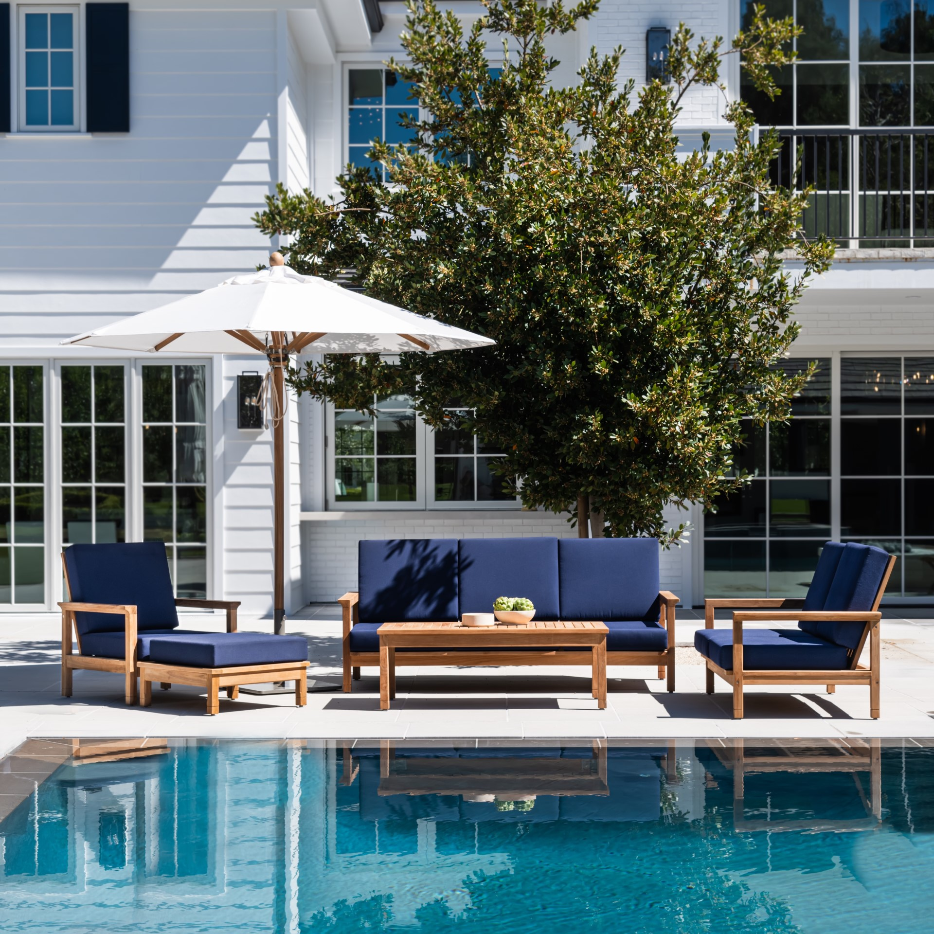 Outdoor furniture set with blue cushions sits under a large umbrella beside a swimming pool. The setting is in a sunny backyard with a white house and lush greenery.