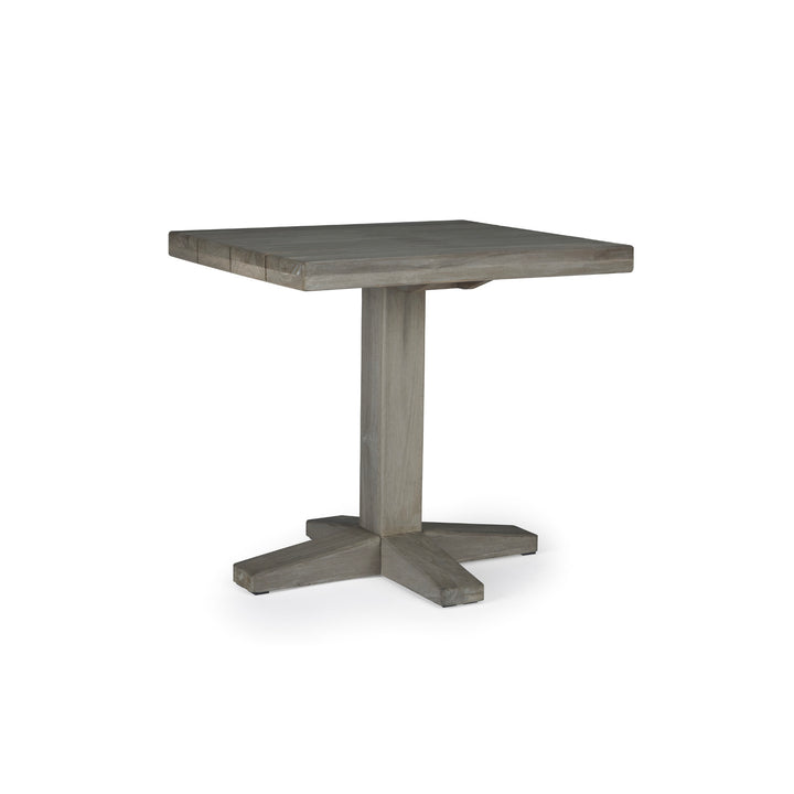 A square wooden table with a central pedestal base stands on a white background. The table has a simple, rustic finish with a cross-shaped support at the bottom.