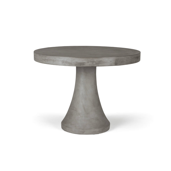 A round, gray concrete table stands on a single, tapered pedestal base in a minimalist, undefined space.