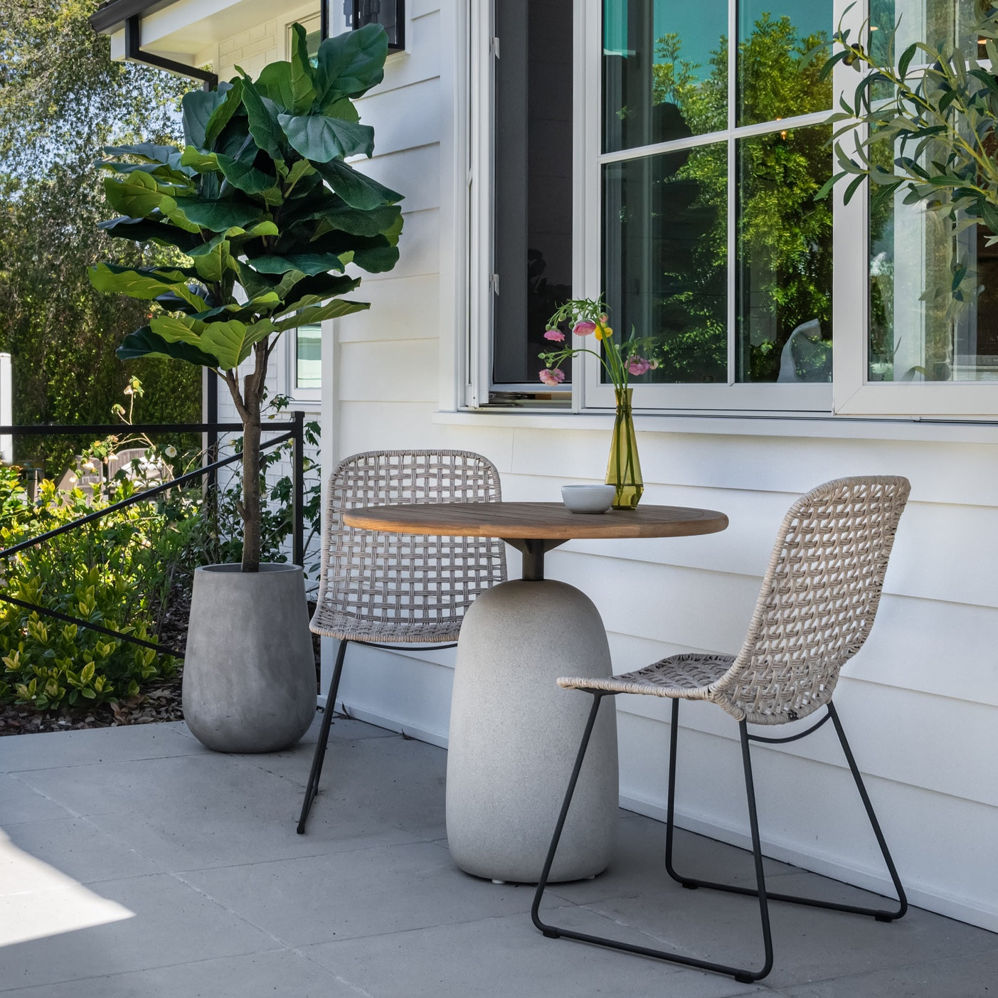 A round wooden table with a vase and cup is flanked by two woven chairs on a patio. Potted plants border the area near a white house with open windows.