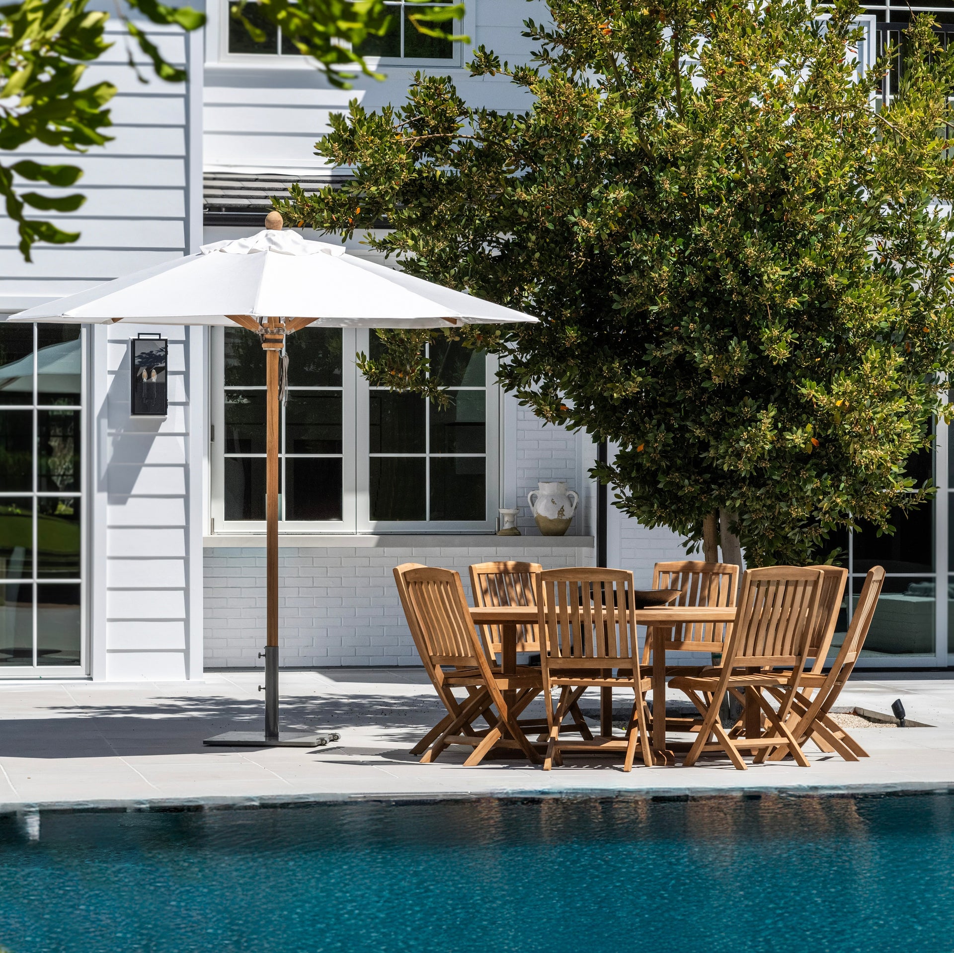 Wooden table and chairs arranged under a white umbrella, situated on a patio beside a swimming pool. The background features a white house and a lush green tree.