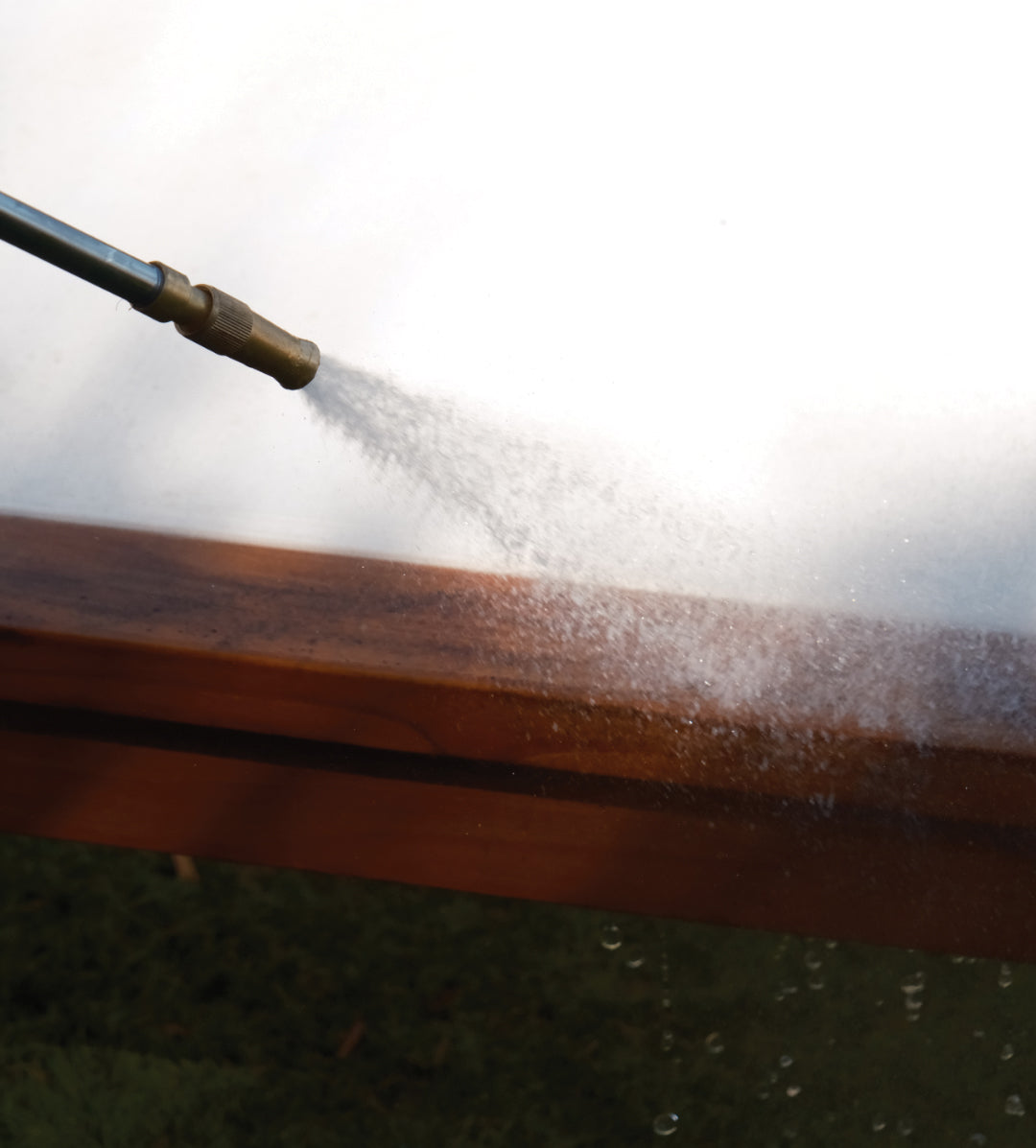 A hose nozzle sprays water onto a wooden surface, creating mist in a bright outdoor setting with grass below.