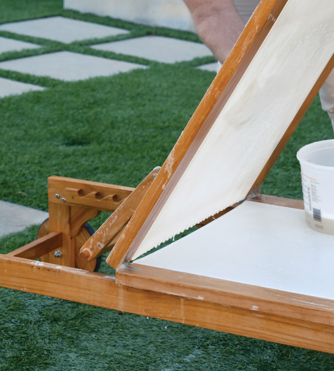 A wooden table with an angled, mounted canvas features a person applying paint outdoors on a grass surface, near a container of paint or plaster.