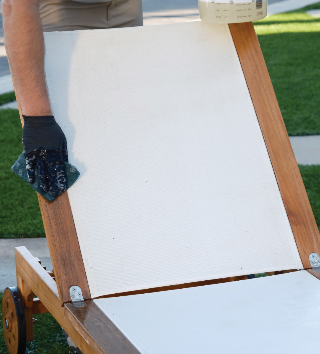 A chaise lounge is being cleaned by a person wearing gloves, using a cloth. The lounge has a white fabric and wooden frame, situated on a lawn with concrete pathways nearby.