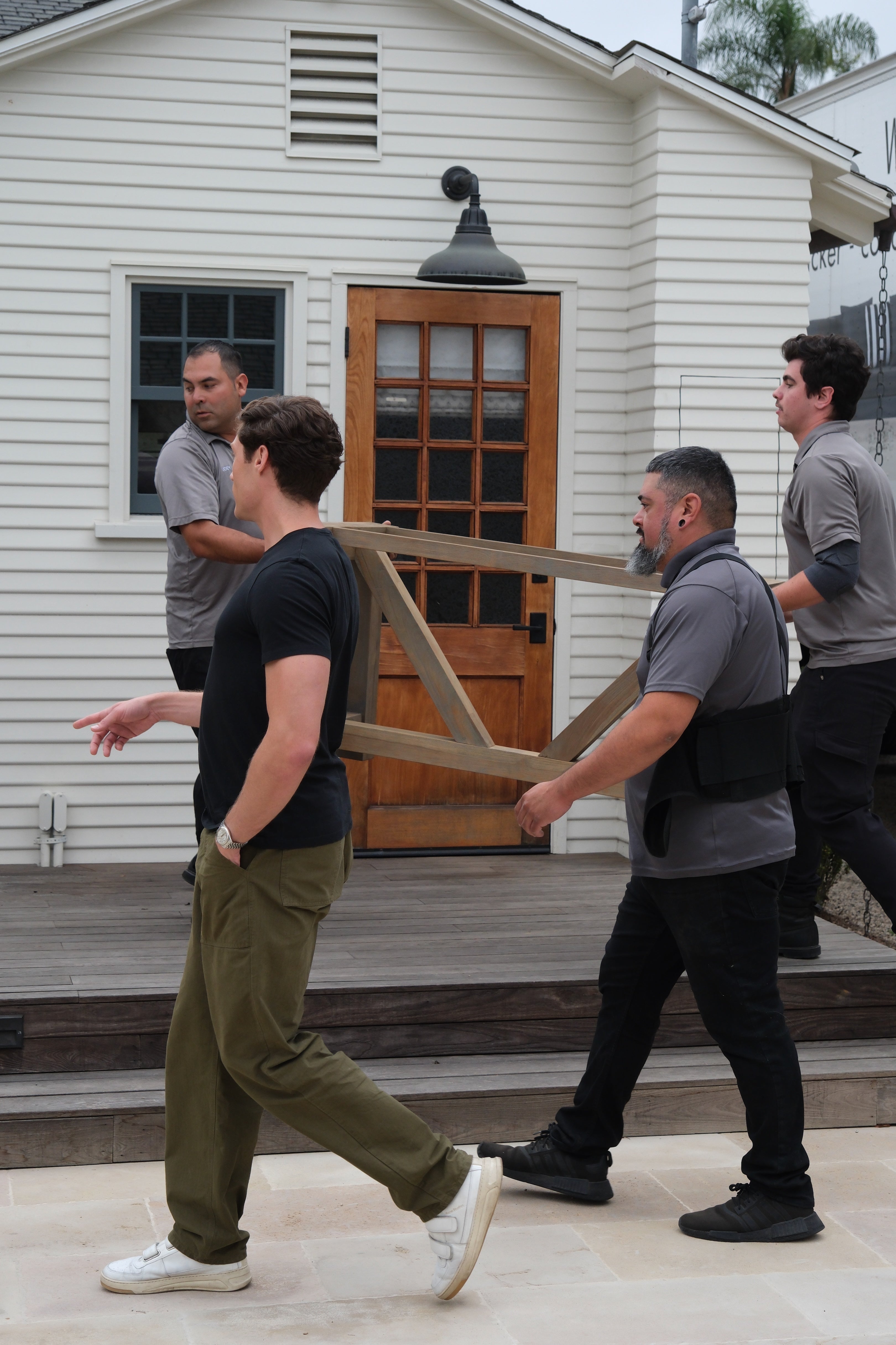 Four men carry a wooden table frame across a wooden deck, in front of a white house with a window and wooden door.