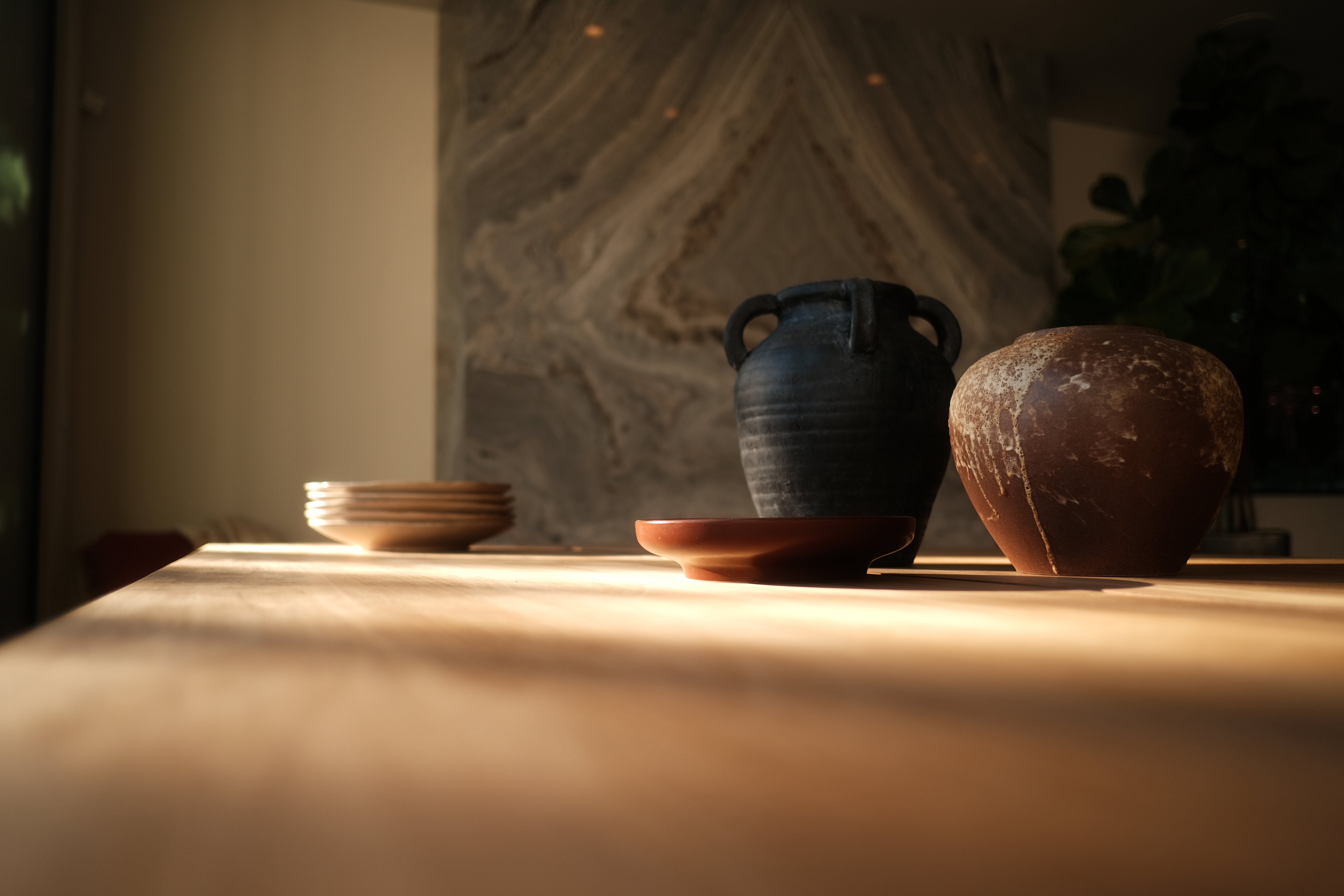 Ceramic pots and stacked plates rest on a sunlit wooden table. The background features a marbled wall and shadows, creating a warm, tranquil atmosphere.