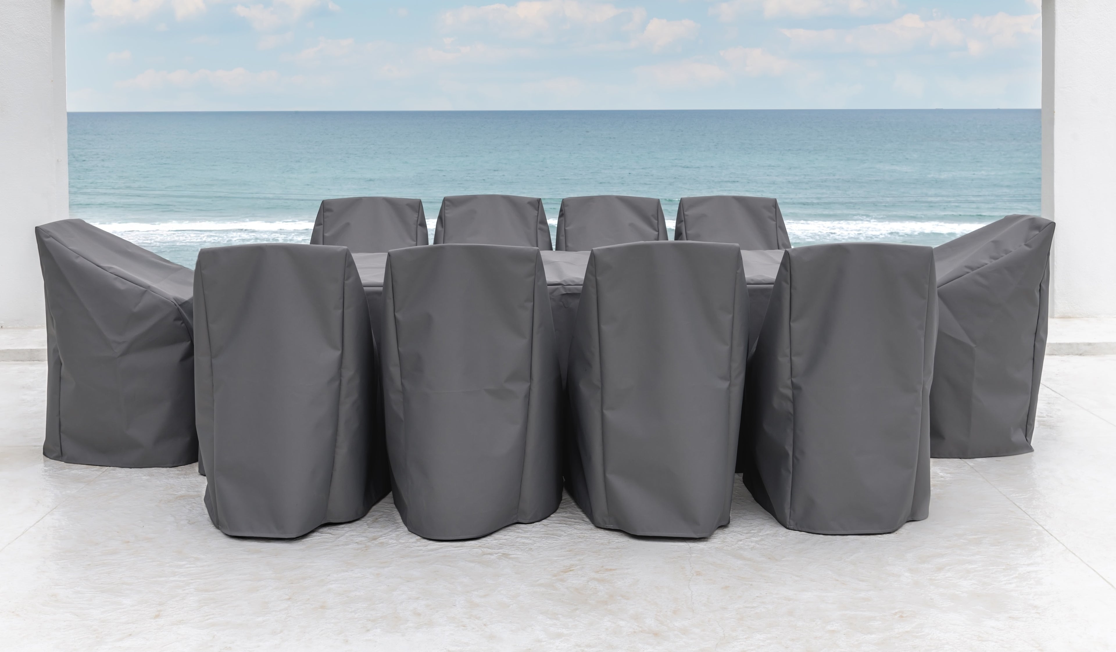 A table and chairs covered with gray protective covers stand on a tiled patio. Behind them, a serene ocean view with a clear sky is visible through two white walls.