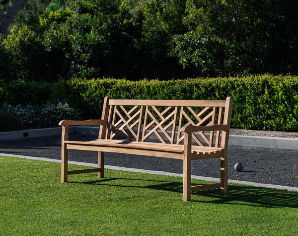 A wooden bench with a geometric backrest design sits on a lawn, surrounded by green hedges and trees, with a g