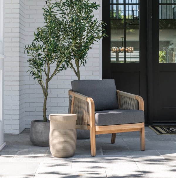 A wooden armchair with dark cushions sits next to a small round table. An olive tree in a pot stands nearby ag